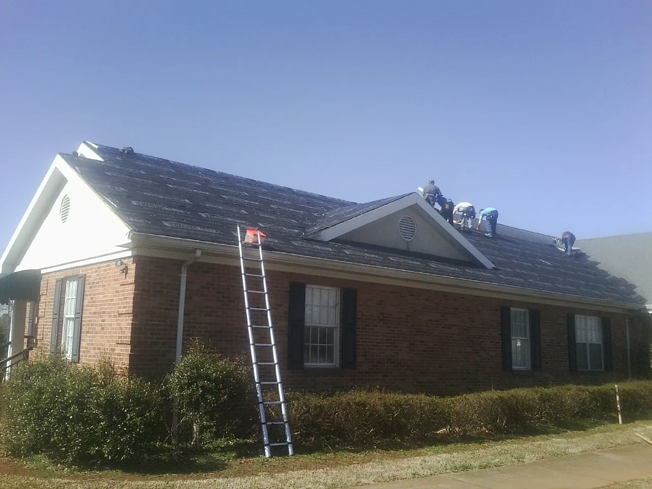 Crew tearing shingles off a brick ranch roof with a ladder set — Emergency Roof Repair work by our crew in Pine