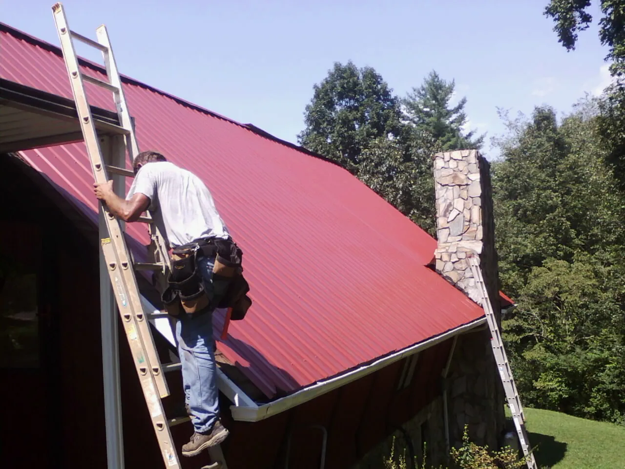 Roofer on a ladder beside a red metal roof and stone chimney — Soffit & Fascia Repair by Roofing Brothers in Pine